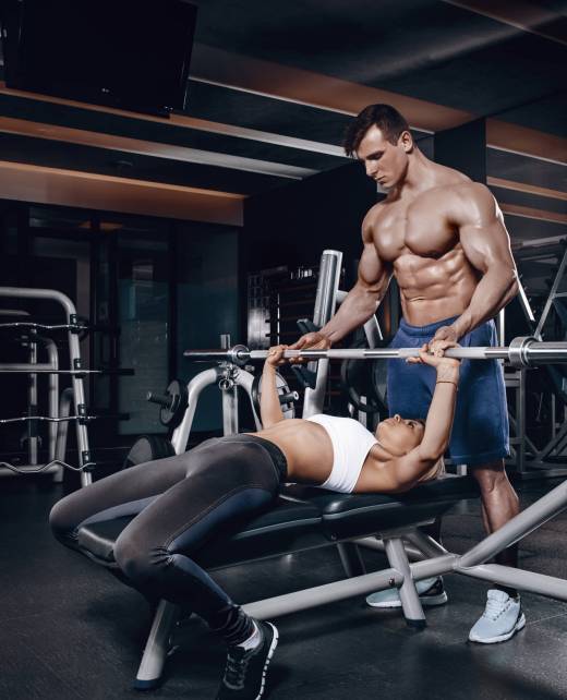 Personal trainer helping a young woman lift a barbell while working out in a gym. Personal trainer with a gorgeous body without a T-shirt in the gym